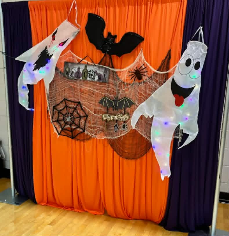Halloween photo booth backdrop featuring orange and purple curtains with hanging ghosts and bat decorations.
