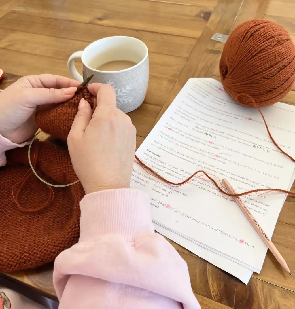 Knitting student following a pattern and knitting with rust coloured yarn on a wooden table