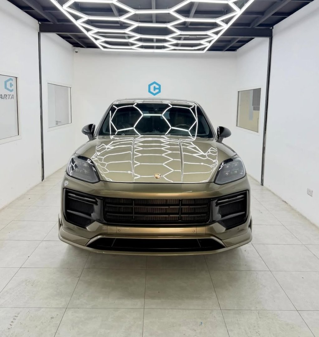 A gold Porsche Cayenne luxury SUV parked in a brightly lit garage with honeycomb LED ceiling lights.