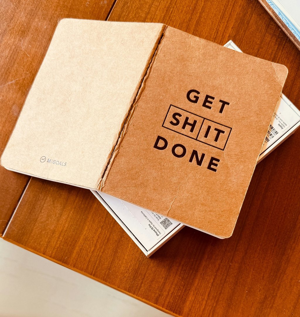 Open MiGoals brown kraft paper notebook with Get Shit Done motivational text on a wooden desk.