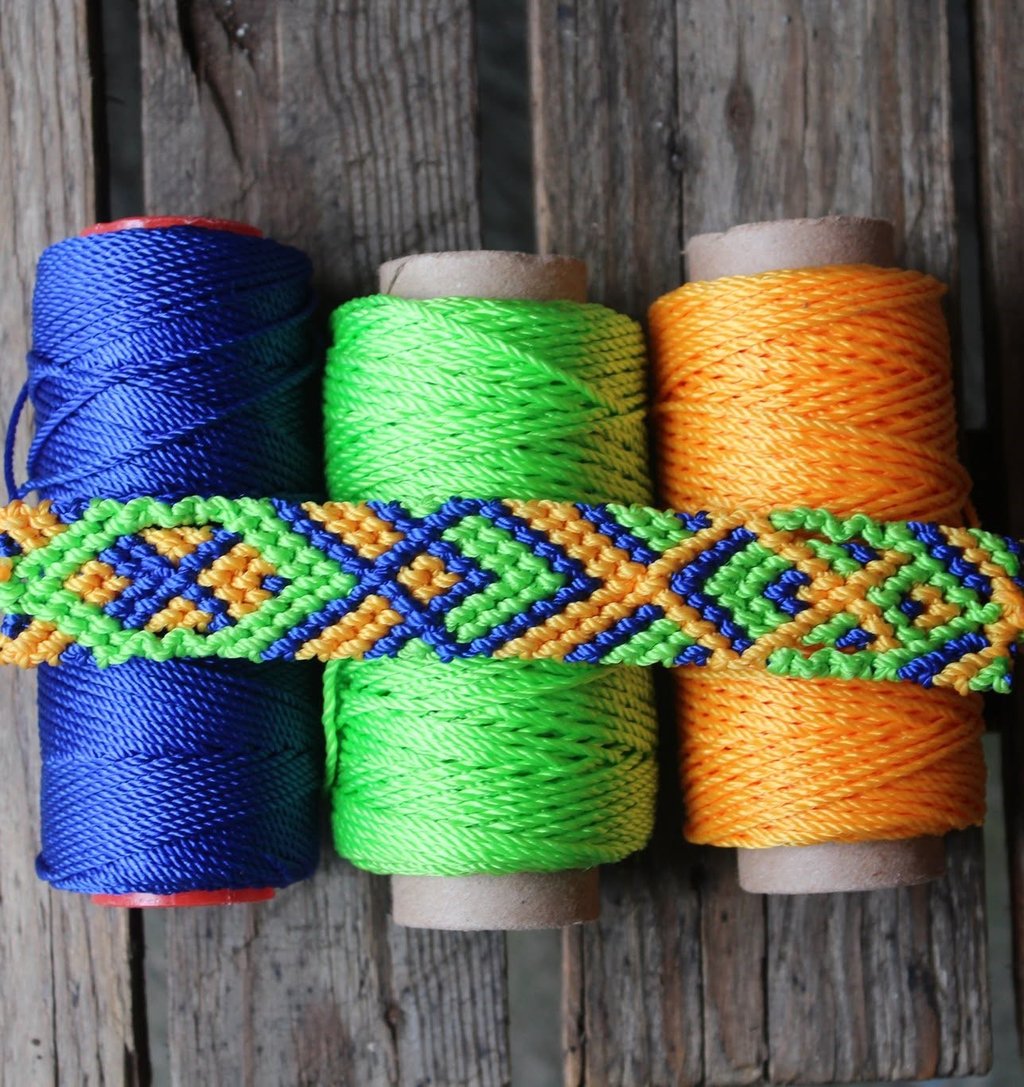 Pulsera  tejida a mano con rombos en azul, verde y naranja, presentada sobre carretes de hilo.
