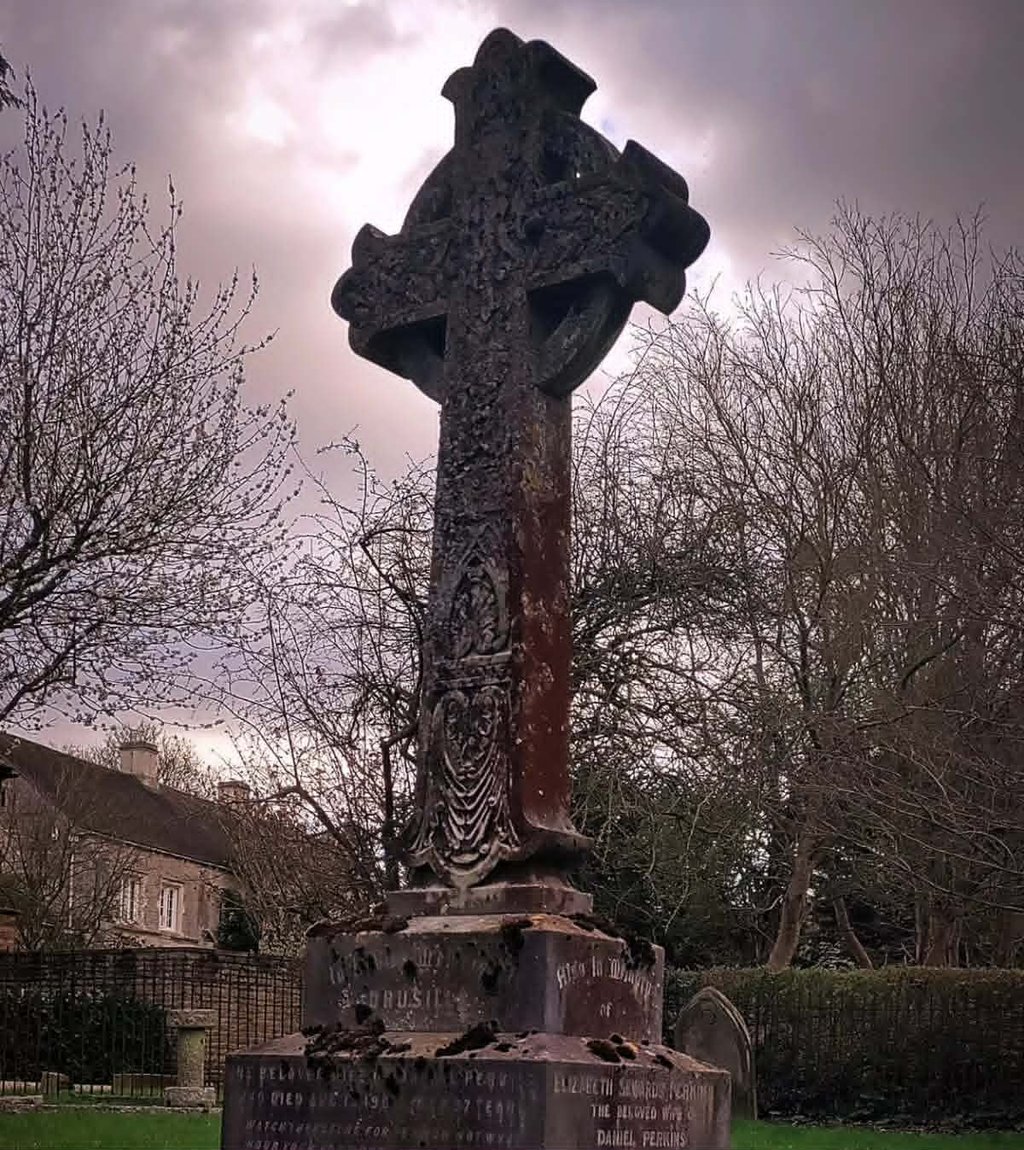 A weathered and lichen-covered Celtic cross headstone in a graveyard with brooding skies overhead