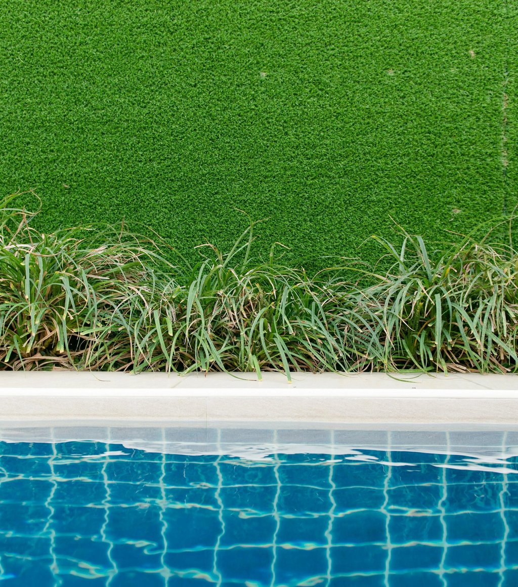 Swimming pool edge with blue water, green plants, and a vibrant artificial grass wall backdrop.