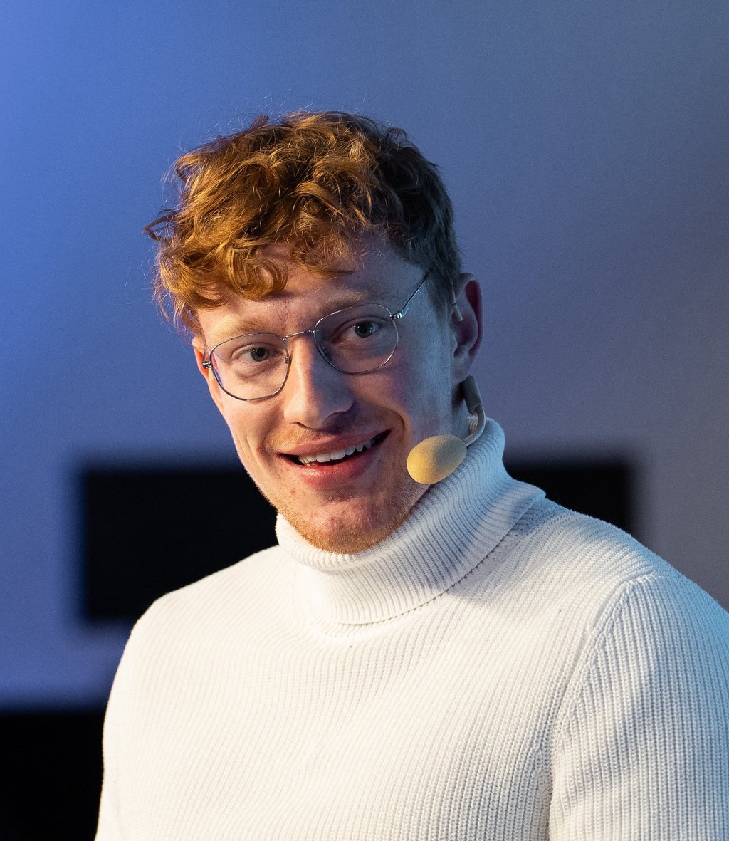 Moderator Przemysław Żuk, kurz Przemek Żuk, moderiert lächelnd eine Paneldiskussion. Er trägt ein Headset-Mikrofon.