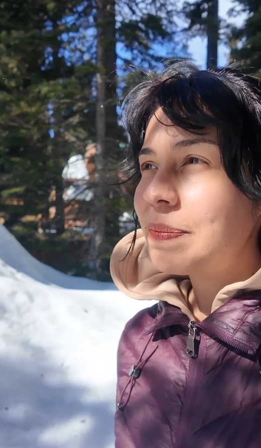 Person standing outdoors in a snowy forest, looking to the side in sunlight with tall trees in the b