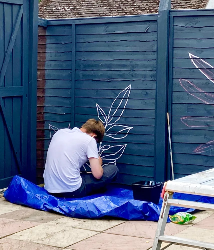 Muralist Byron sits on floor, painting graphic plant on grey wooden garden fence.