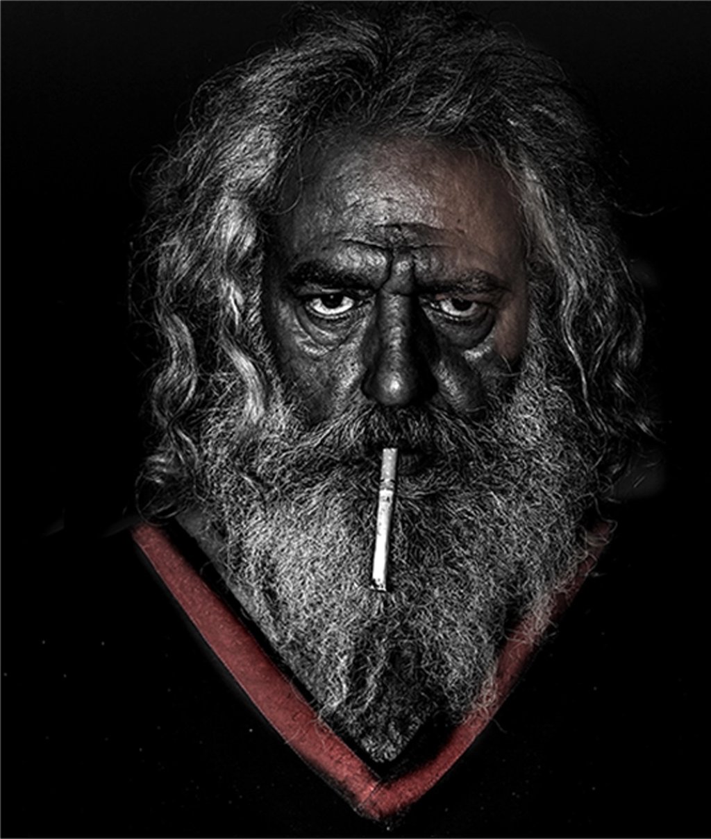 Portrait of an older man with a weathered face and intense gaze, cigarette held between his lips. 
