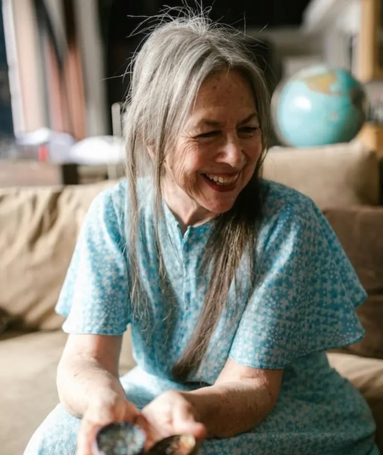 Smiling senior woman with long grey hair wearing a blue floral dress while sitting on a sofa.