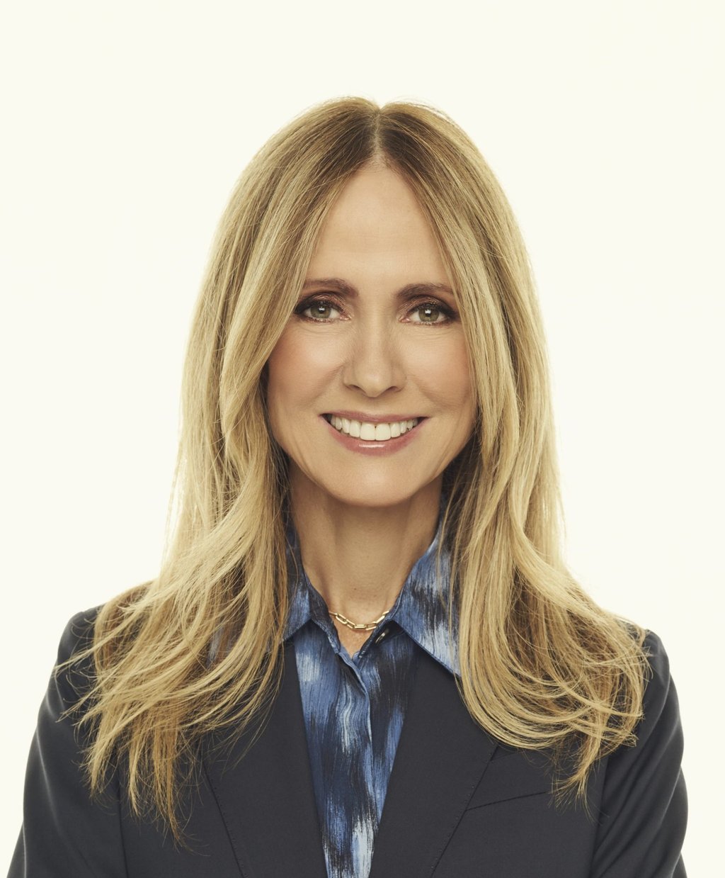 Professional headshot of a smiling blonde woman in a navy blazer and blue patterned shirt.