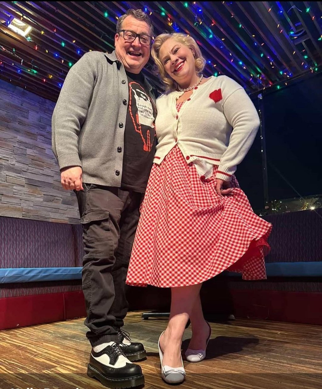 A smiling couple in retro-style clothing poses under colorful lights at a rockabilly event.