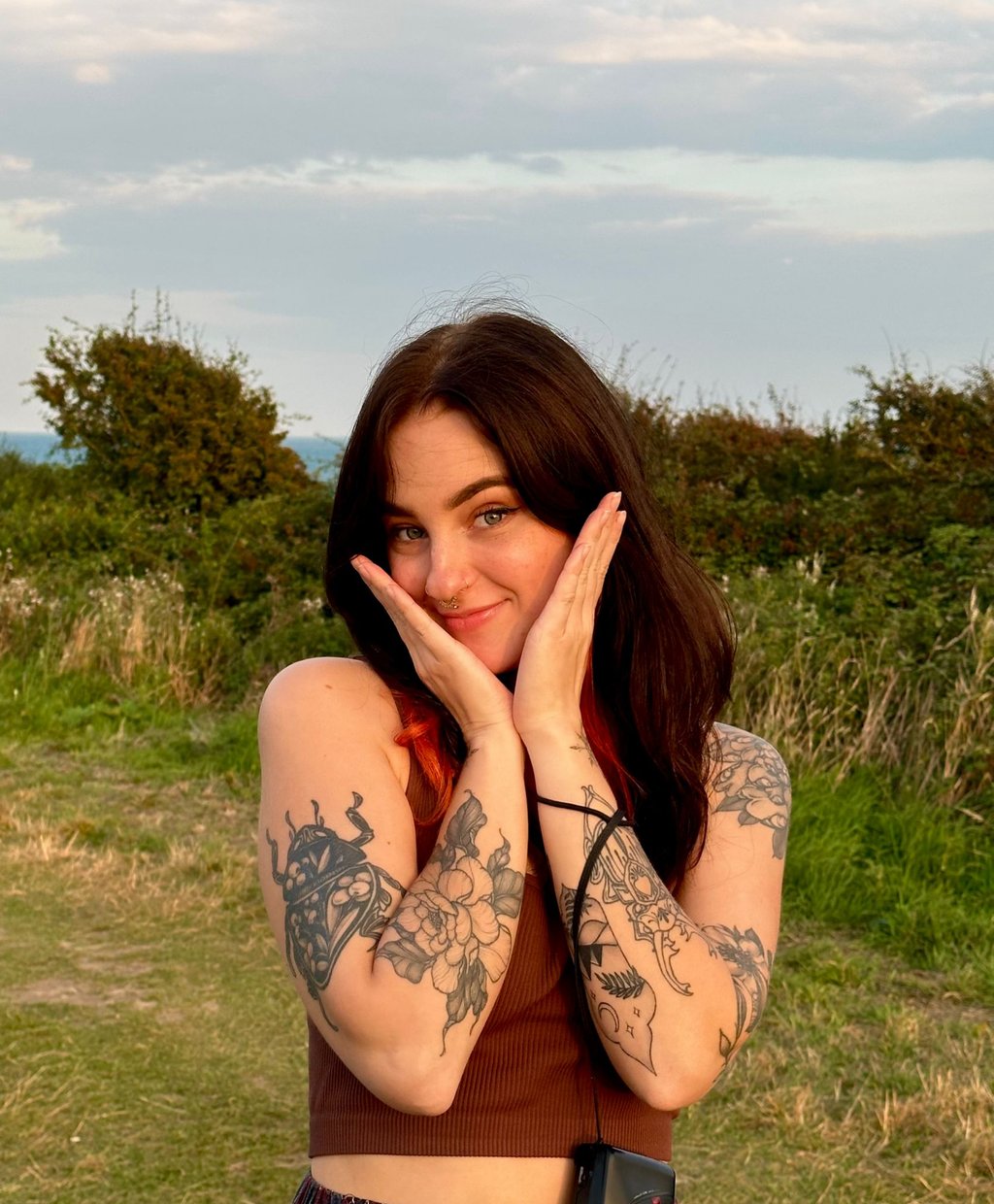 Smiling woman with dark hair and floral arm tattoos posing outdoors in nature at sunset.