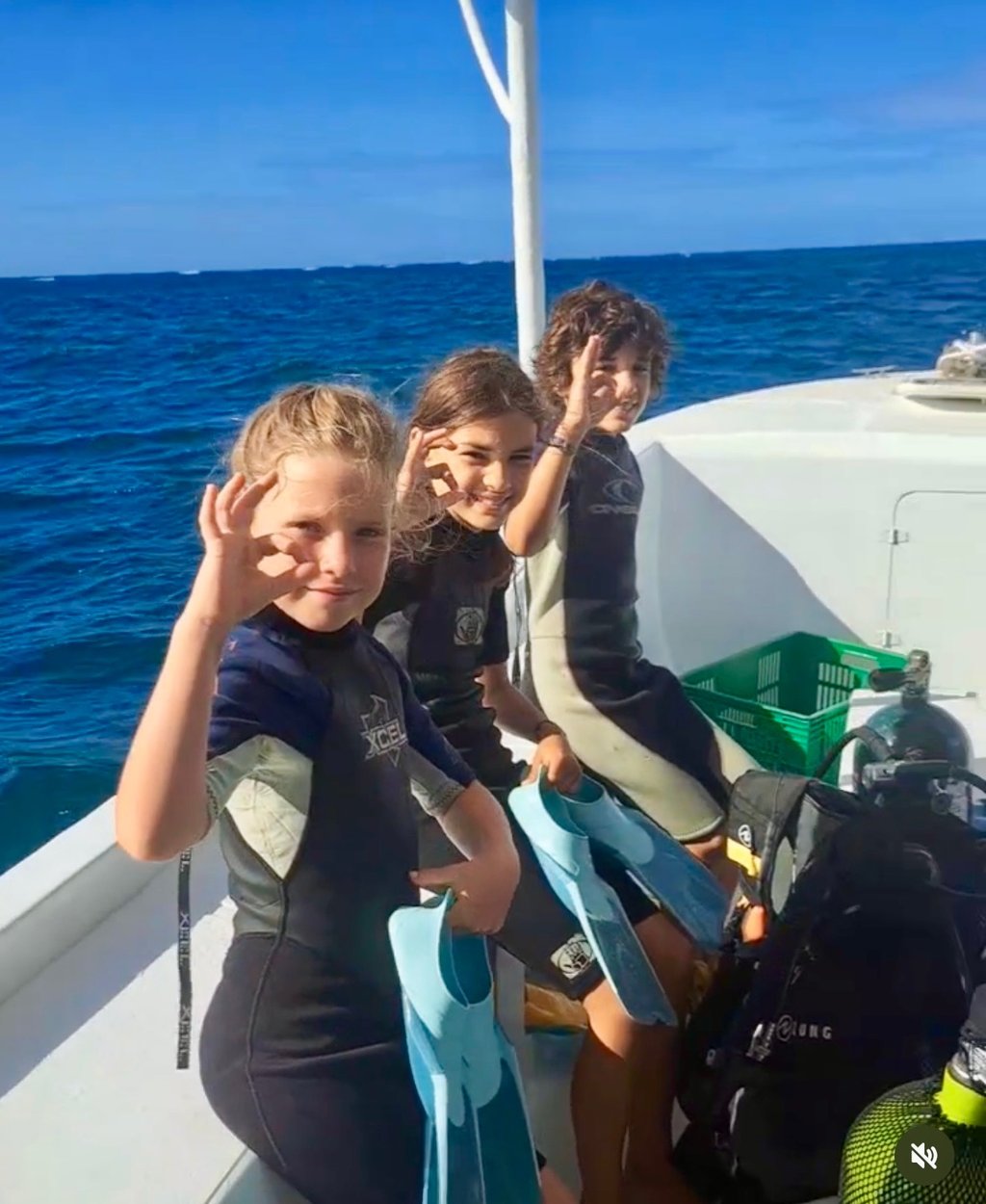 Three children in wetsuits give the OK sign on a dive boat while holding flippers for snorkeling.