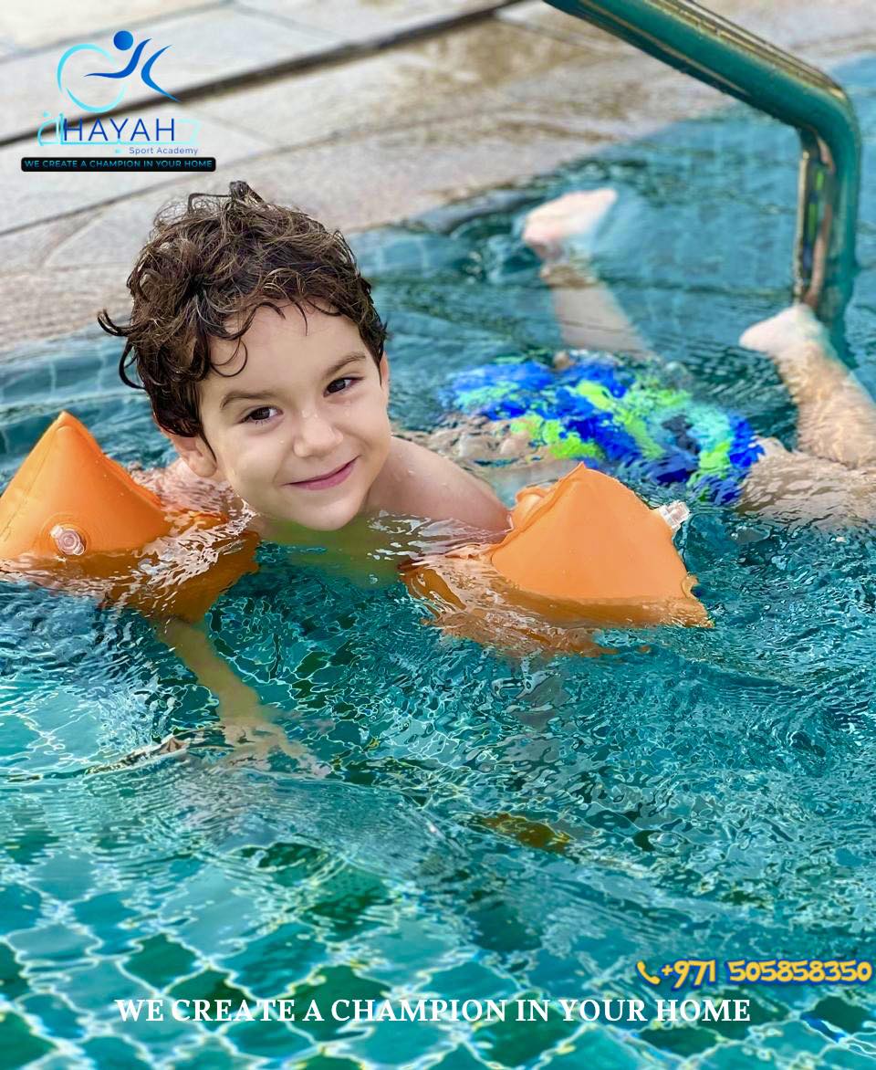 A smiling young boy with orange floaties learns to swim in a pool during Hayah Sport Academy lessons.
