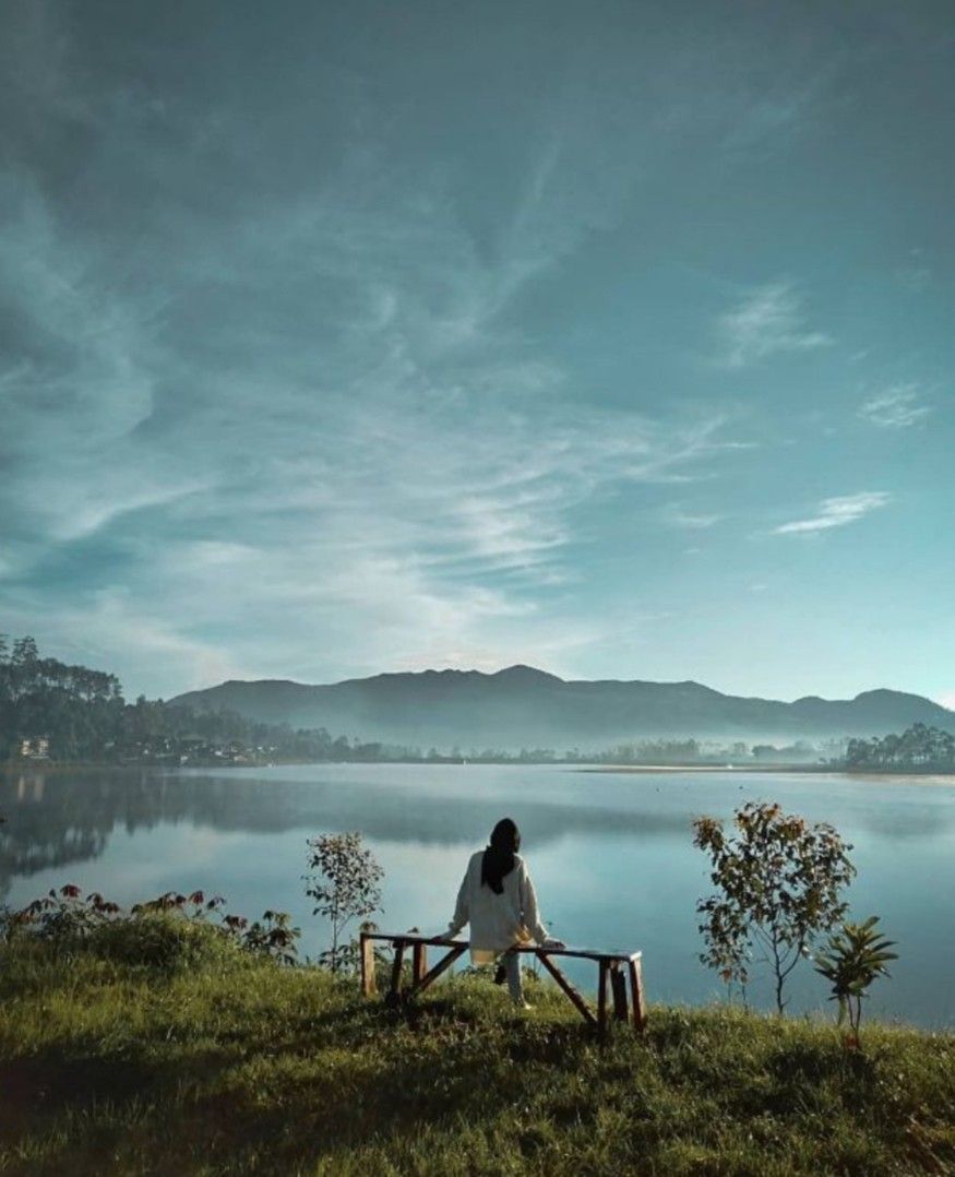 Seorang wanita berhijab duduk di bangku kayu menghadap danau Situ Cileunca yang tenang dengan latar belakang pegunungan dan l