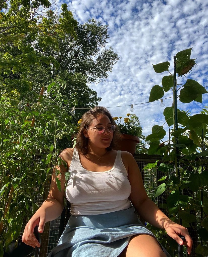 A woman wearing glasses and a white tank top sits in a lush green garden under a blue sky.