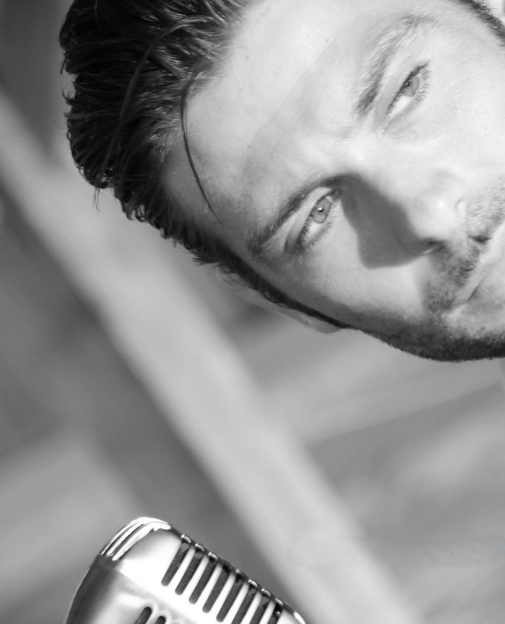 Black and white photo of a handsome man with slicked-back hair looking down at a vintage microphone, perhaps a singer or