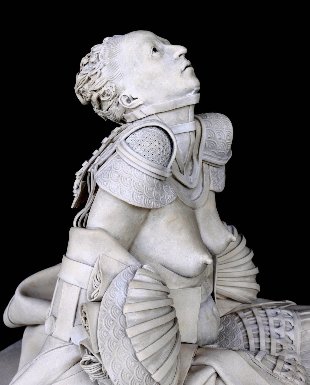 Ceramic sculpture of fallen nude woman in samurai skirt, shoulder armor and neck brace pleading upward; closeup right side..