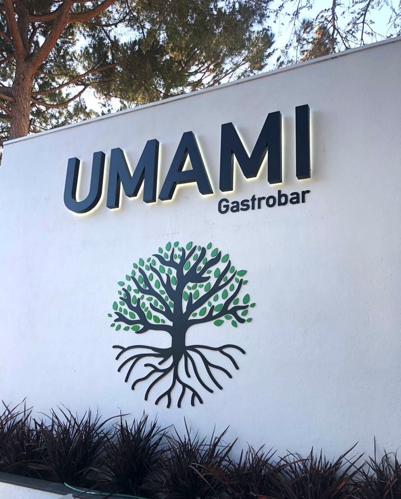 Umami Gastrobar signage on a white wall with a decorative tree logo and outdoor landscaping.