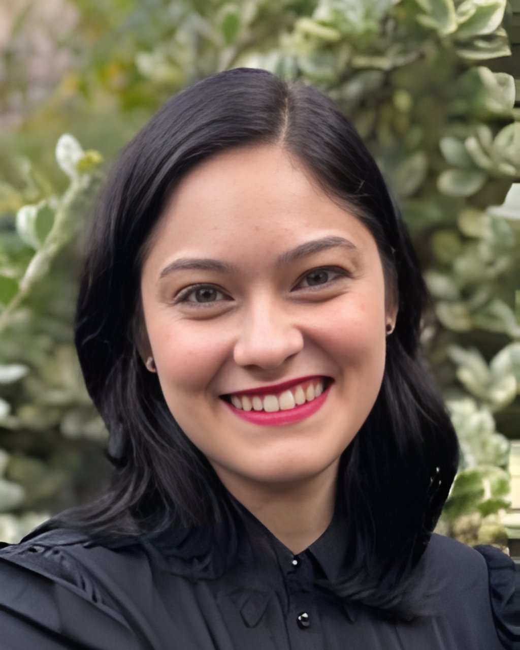 Smiling woman with black hair and red lipstick posing against a blurred green garden background.