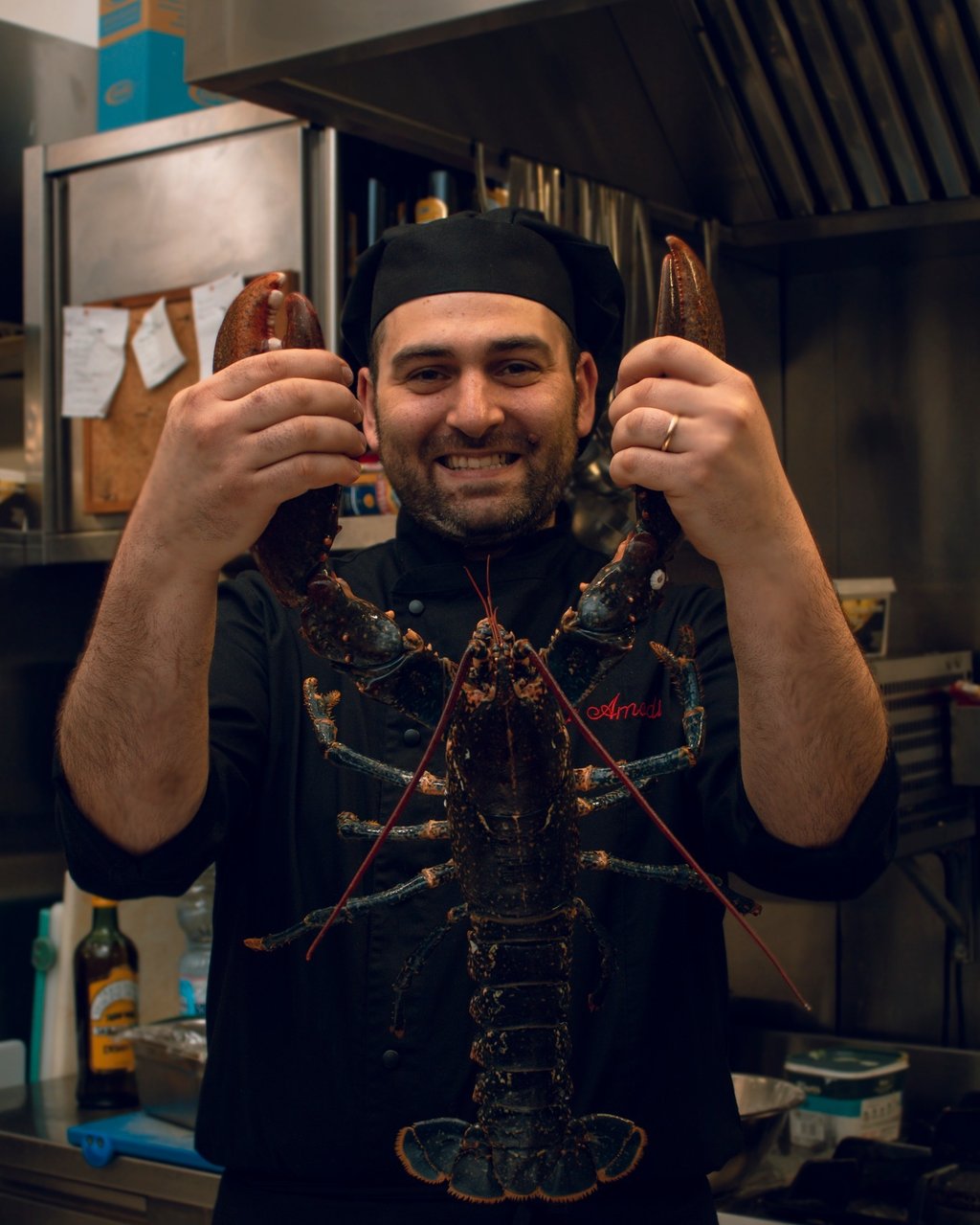 Chef Vito Amodio in cucina con divisa, sorridente mentre tiene in mano un grande astice vivo