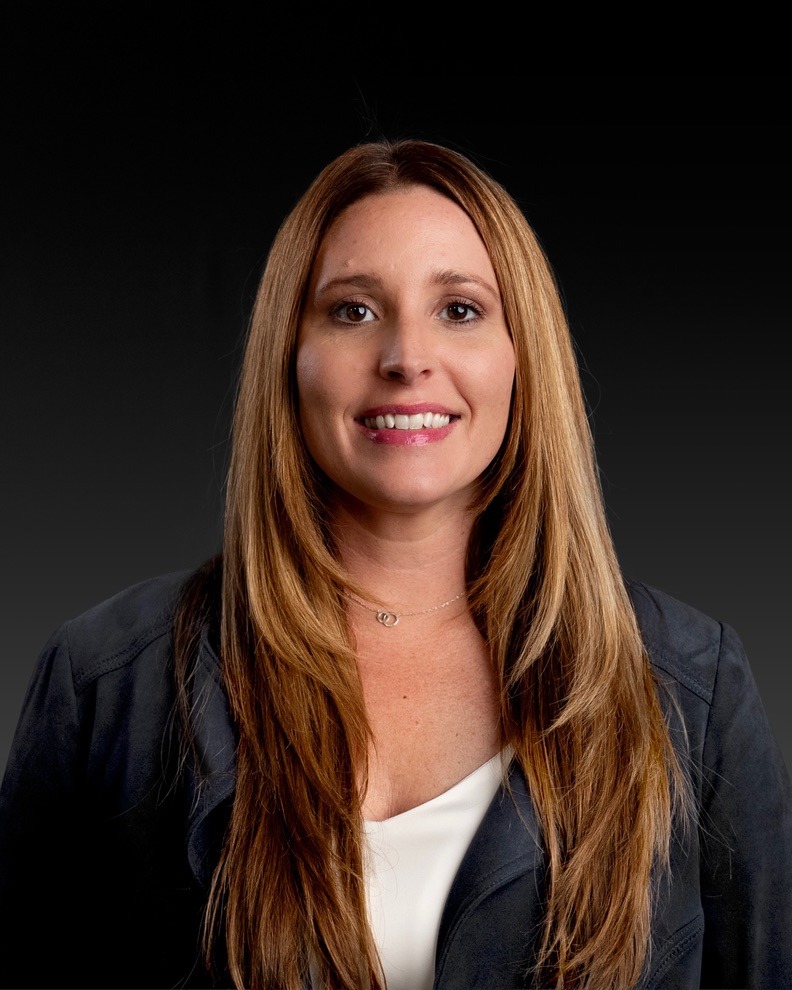 Professional corporate headshot of a smiling Tara Follett with long brown hair wearing a blazer.