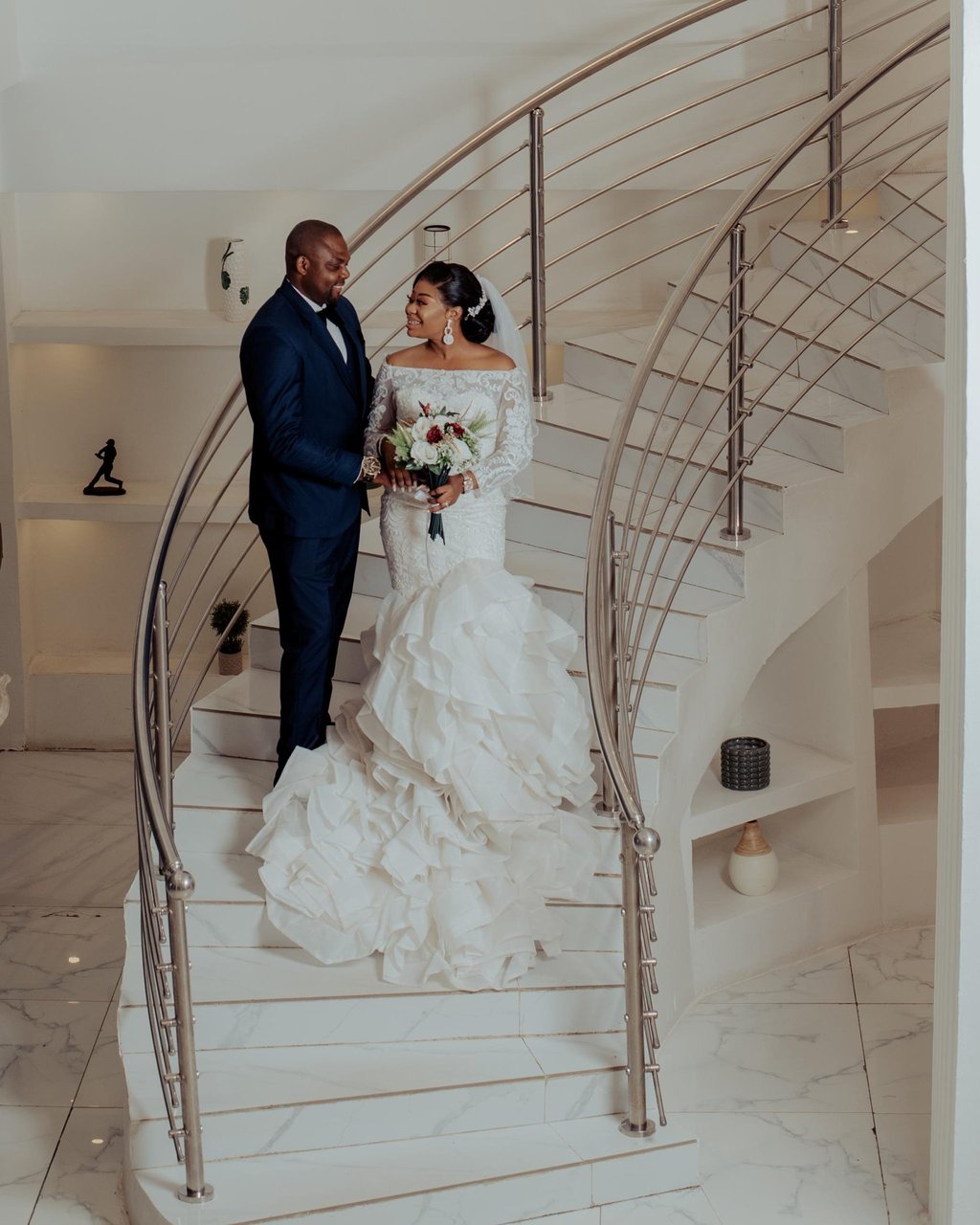 Smiling bride in a ruffled mermaid wedding dress and groom in a navy suit on a white marble spiral staircase.