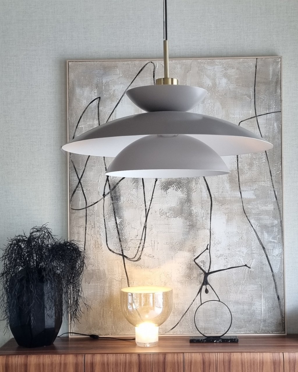 Modern grey tiered pendant light hanging above a buffet with abstract art and a glowing glass table lamp.