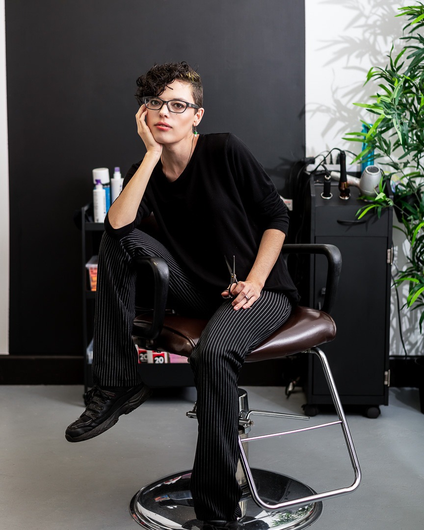 A hairdresser sits in a salon chair holding posing with a pair of scissors