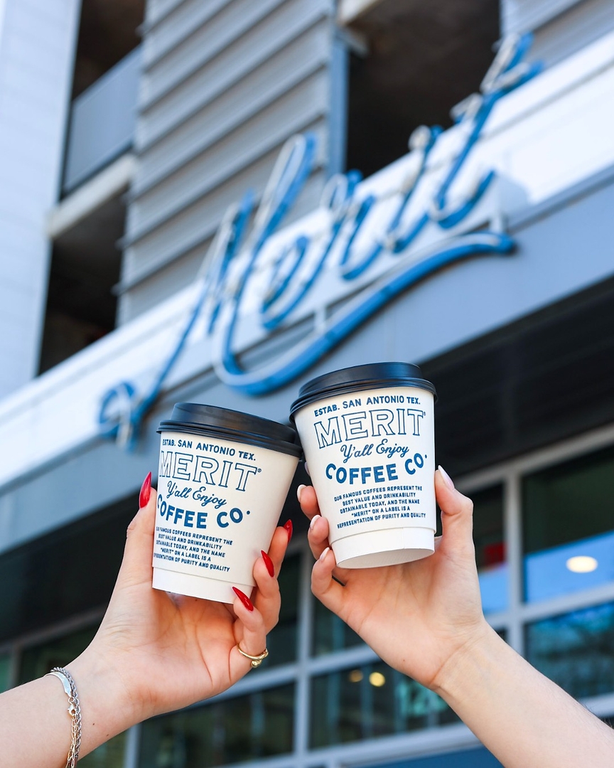 Two hands holding Merit Coffee Co. cups in front of a modern San Antonio cafe storefront.