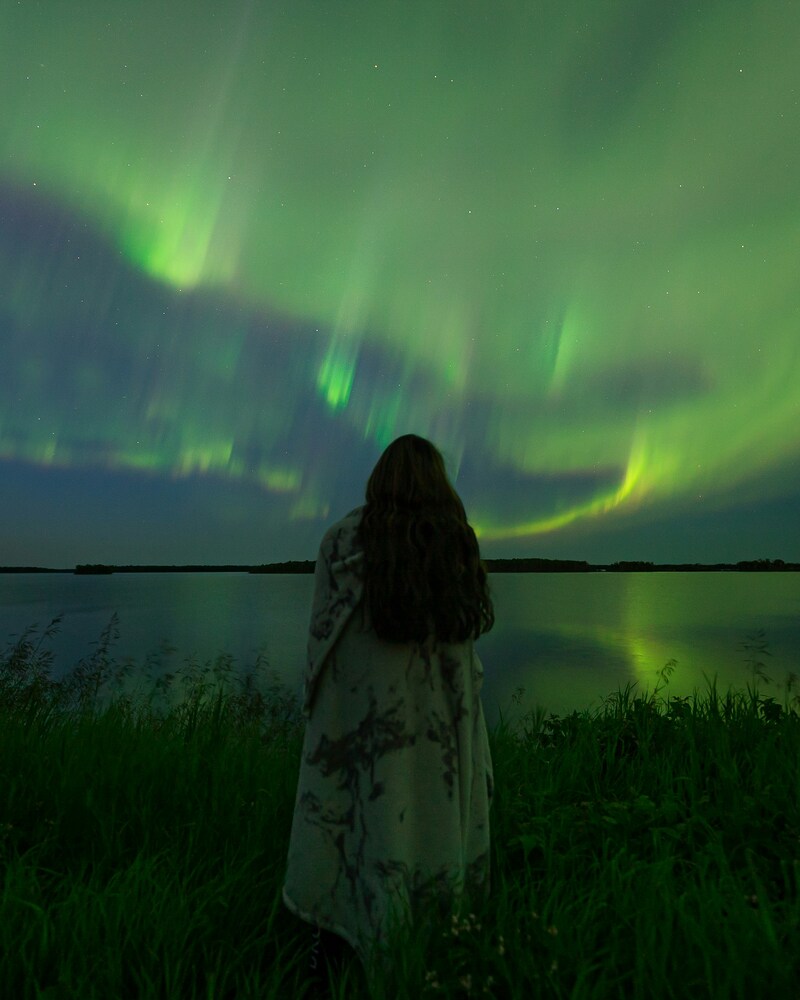 A person watching the vibrant green Aurora Borealis northern lights reflected over a calm lake at night.