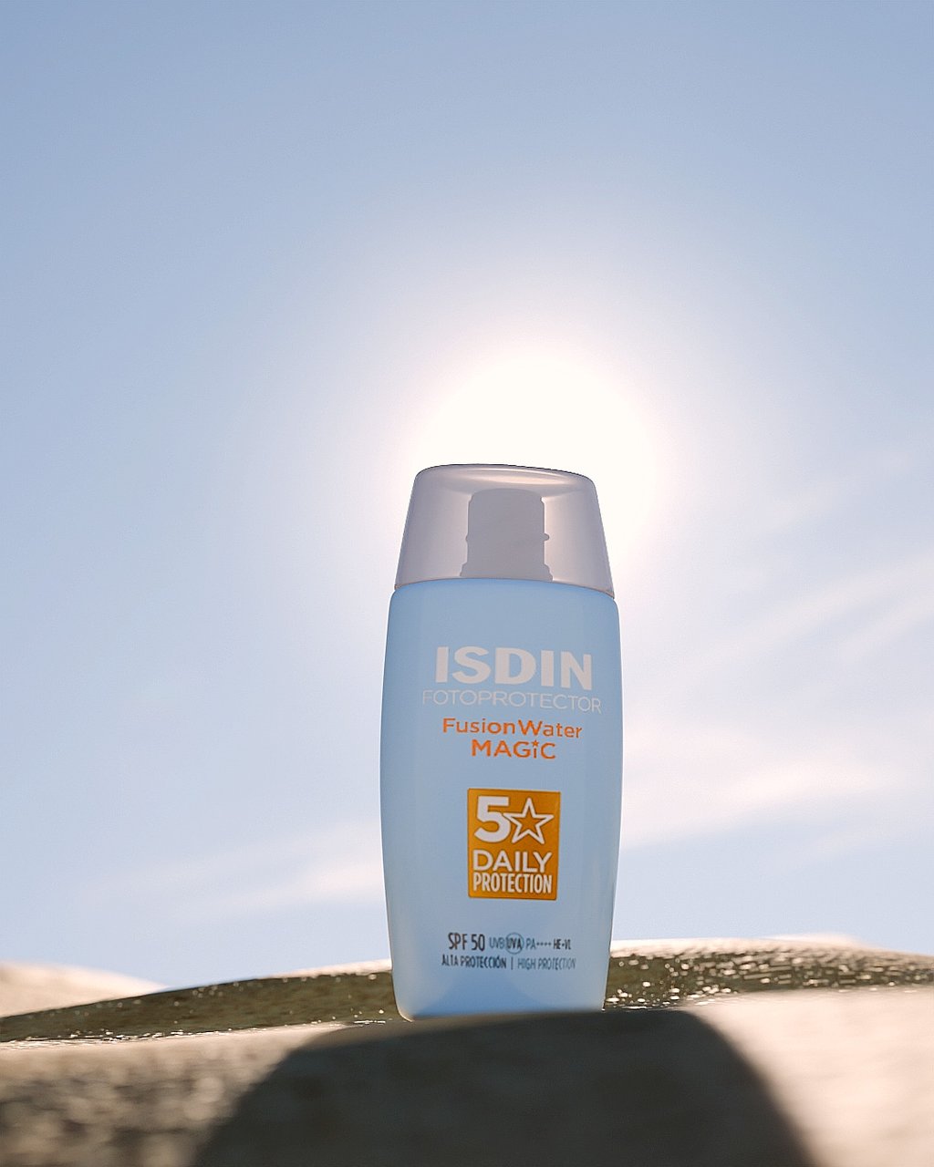 Bote de crema de sol de ISDIN en la arena y el sol detrás.