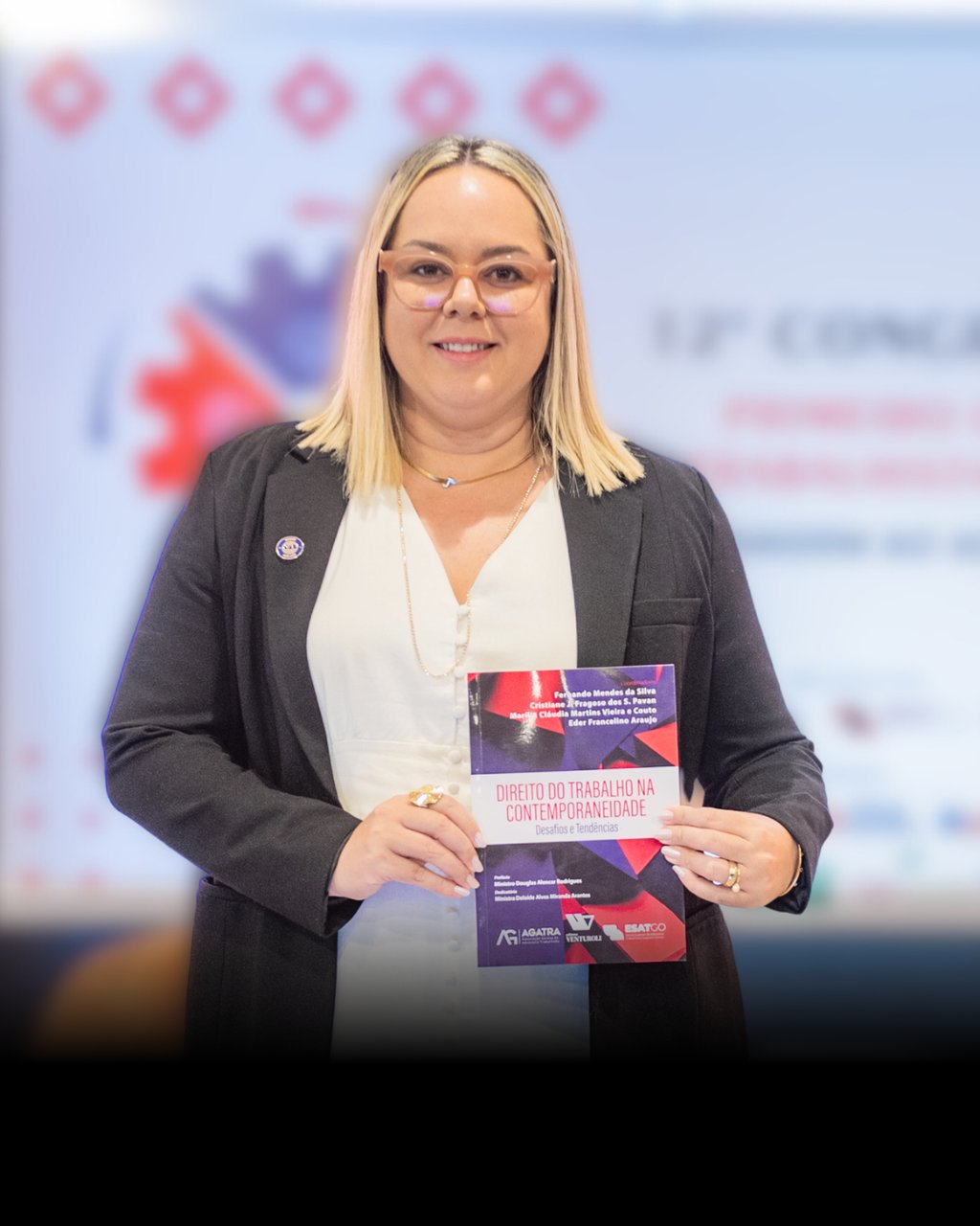 Uma mulher loira de terno e óculos segurando um livro.