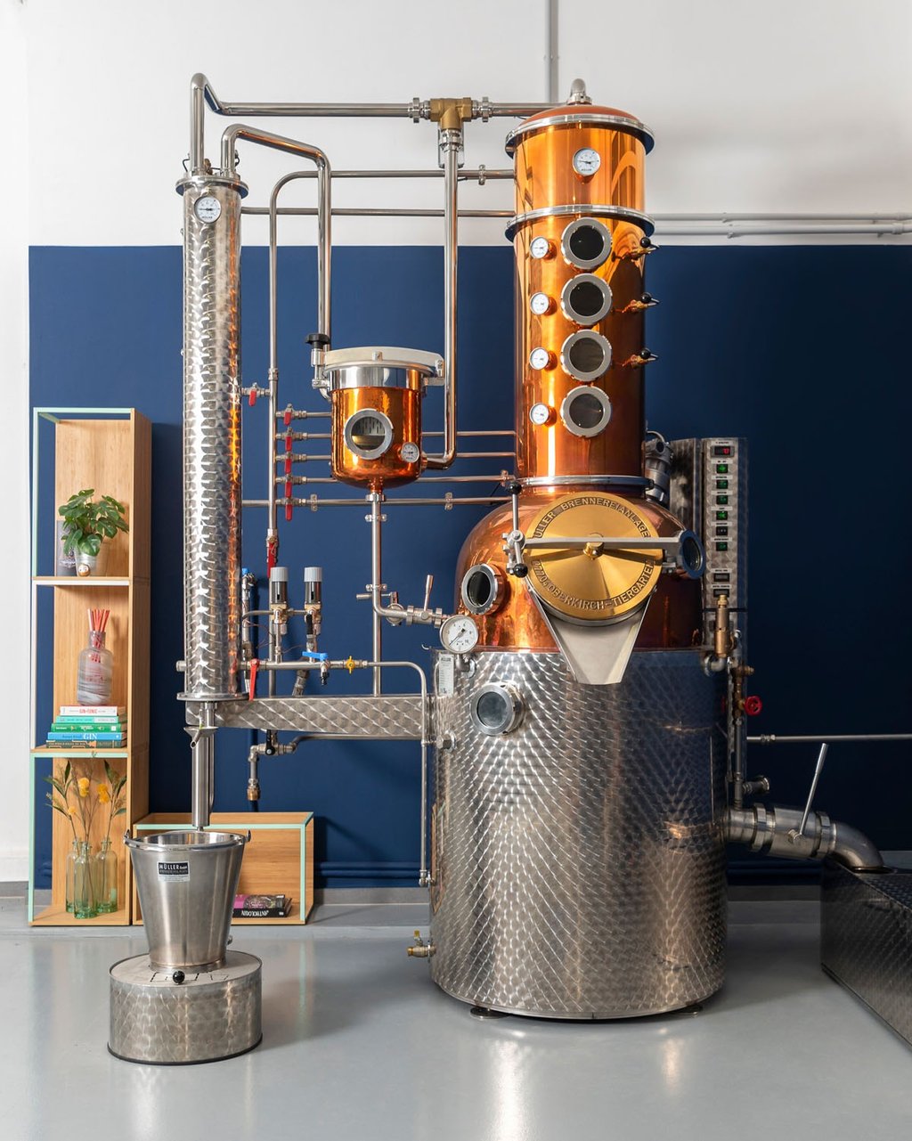 un bellissimo alambicco in rame, acciaio e ottone con un mobile, piante e libri su una parete blu