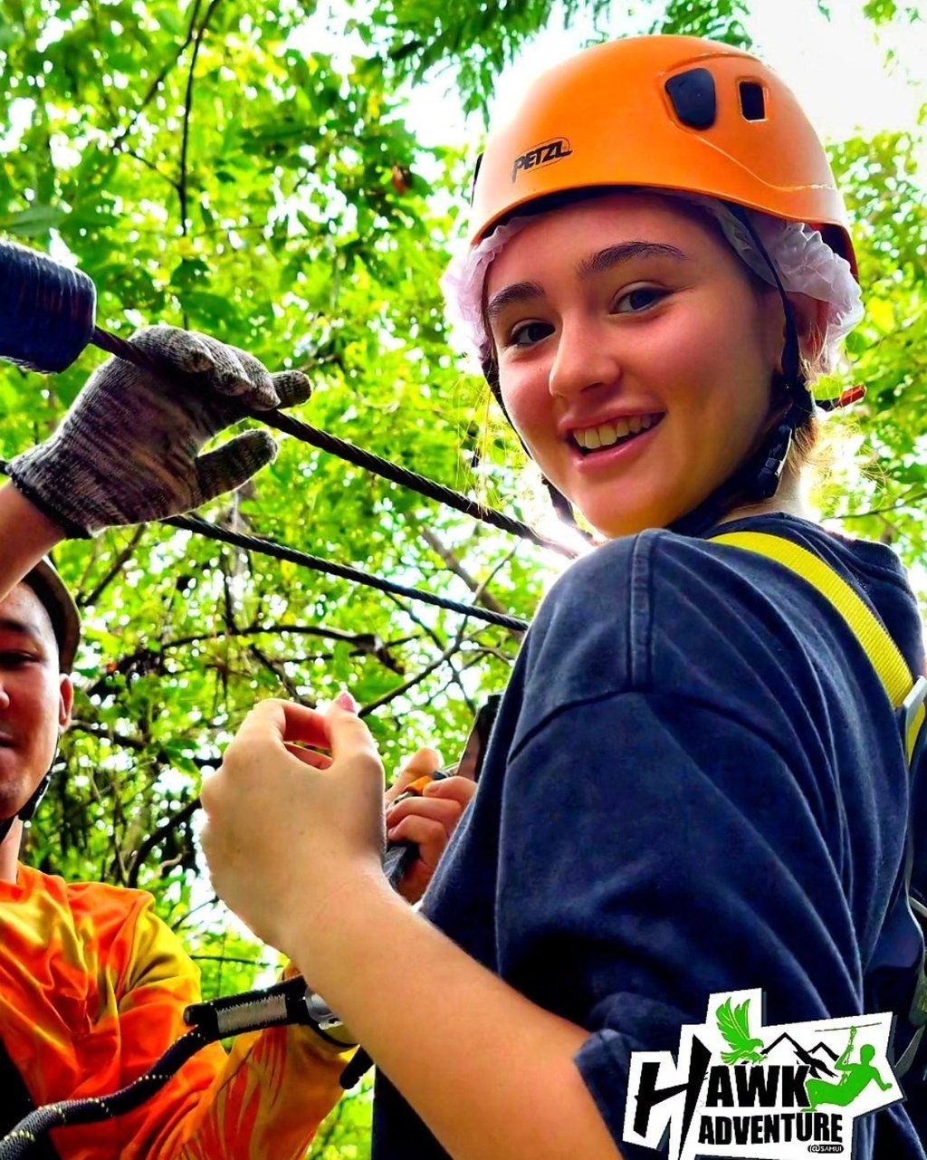 Smiling girl wearing an orange Petzl helmet and safety harness at a Hawk Adventure ropes course.