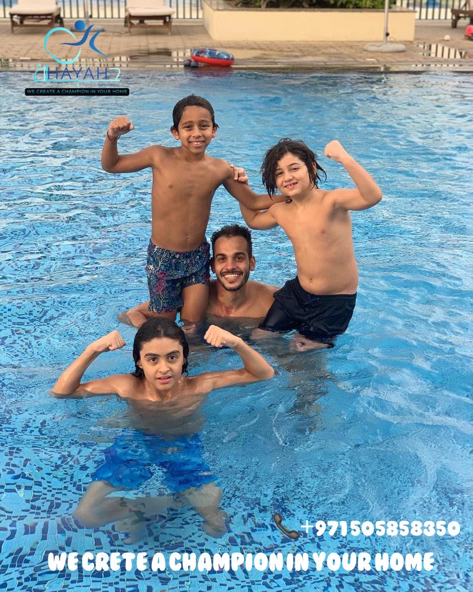A group of boys and their swimming coach smiling and flexing muscles in an outdoor swimming pool.