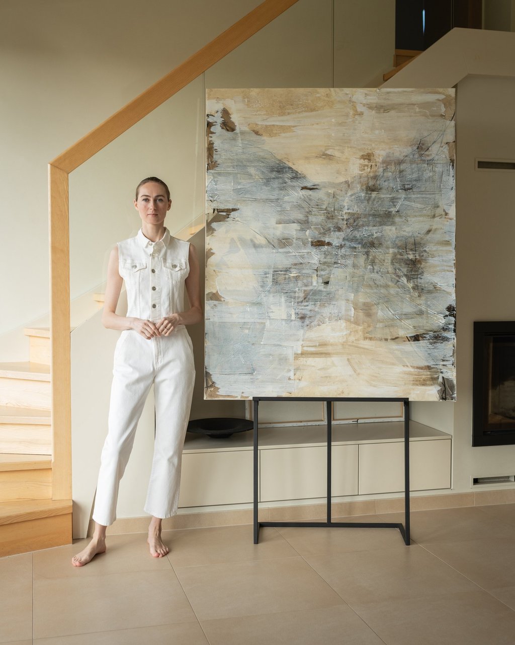 A female artist in a white jumpsuit stands by a large abstract painting in a modern home.