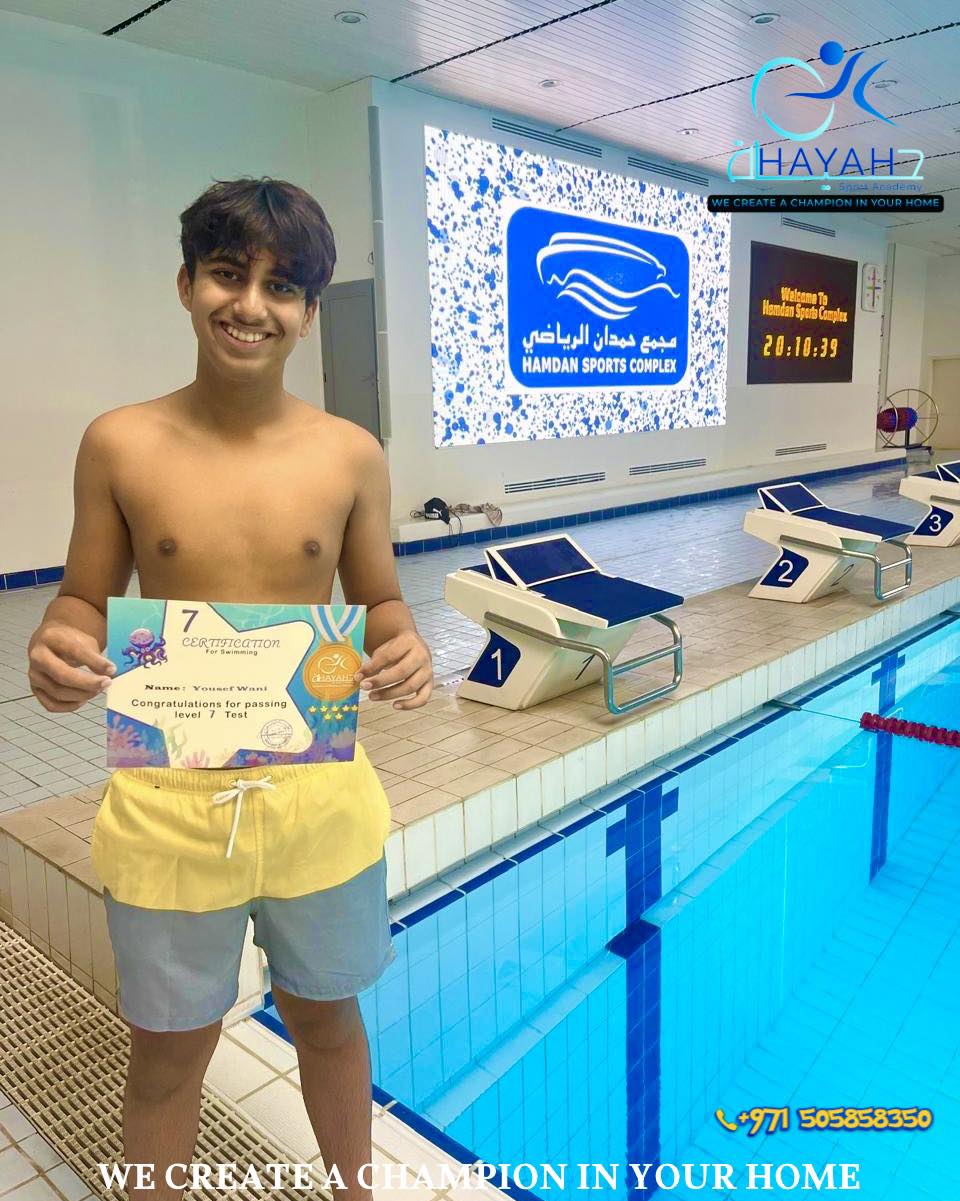 A young male swimmer holding his Level 7 swimming certificate at Hamdan Sports Complex pool.