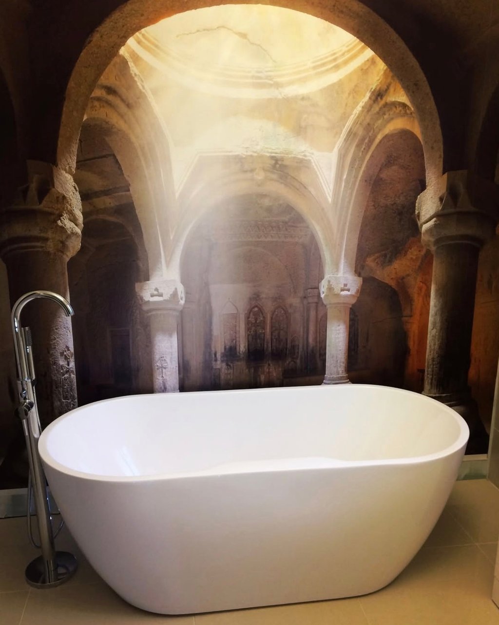 Modern white freestanding bathtub set against a mural of an ancient stone cathedral with sunlight.