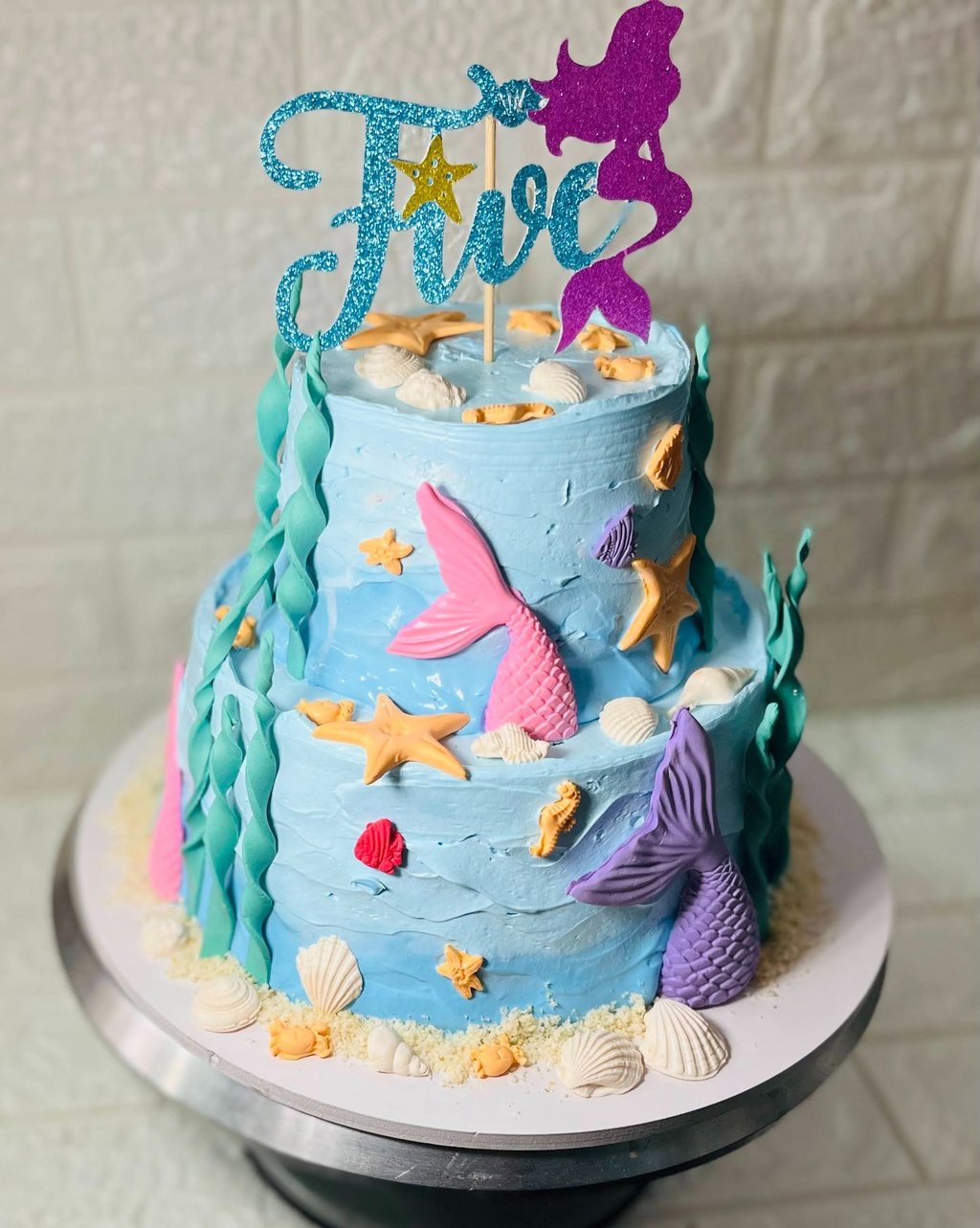 Two-tier light blue mermaid birthday cake for a fifth birthday decorated with shells and tails.