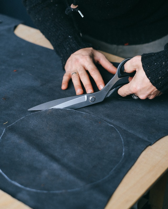 Hands cutting a circular fabric patch from dark denim using large fabric scissors.