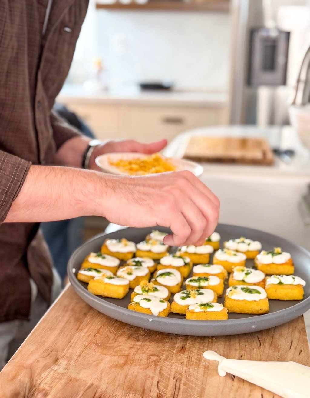 A chef garnishes a tray of crispy polenta squares topped with creamy goat cheese and fresh herbs.