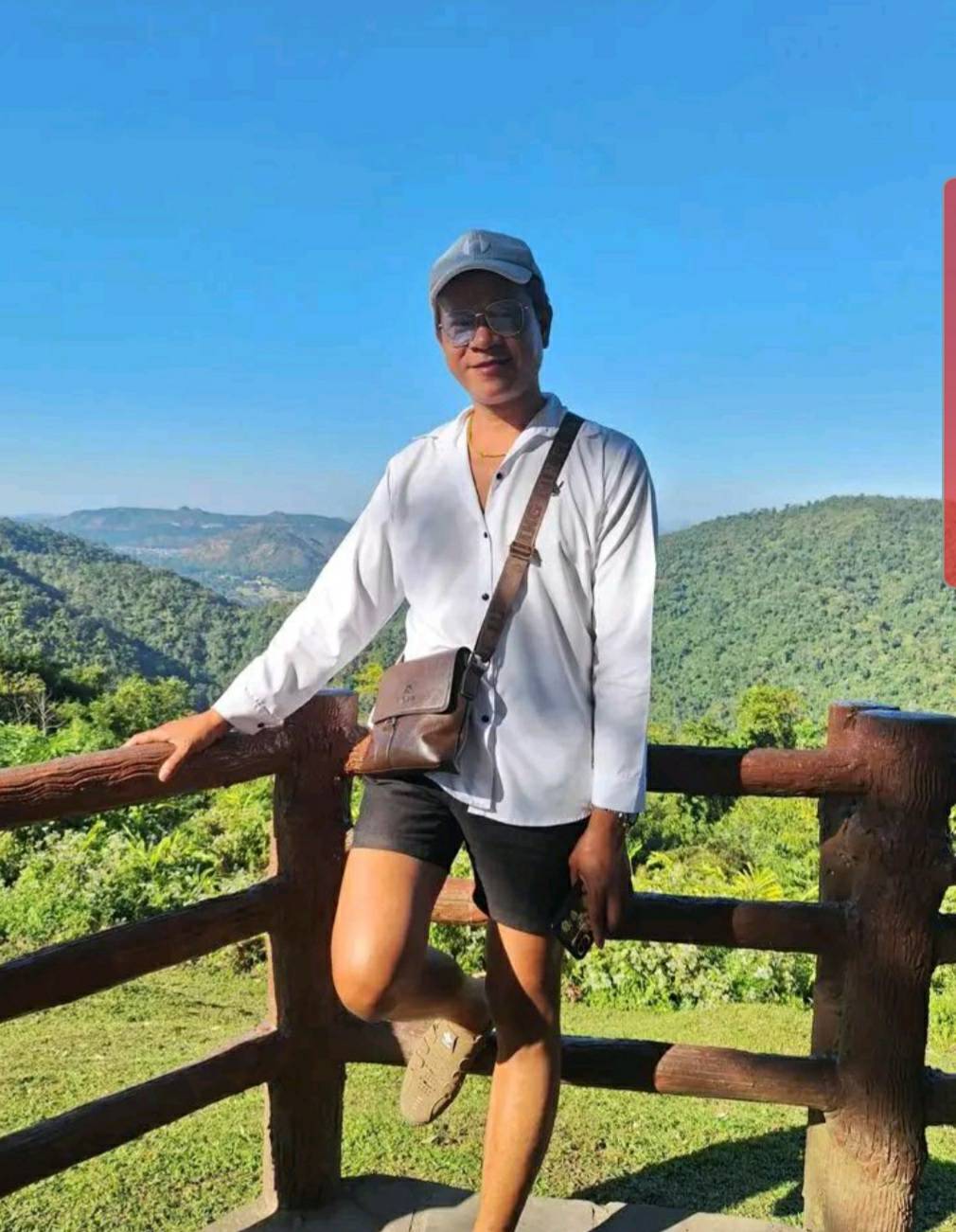 Man standing by a wooden railing overlooking green hills and blue sky in Pattaya, Thailand.