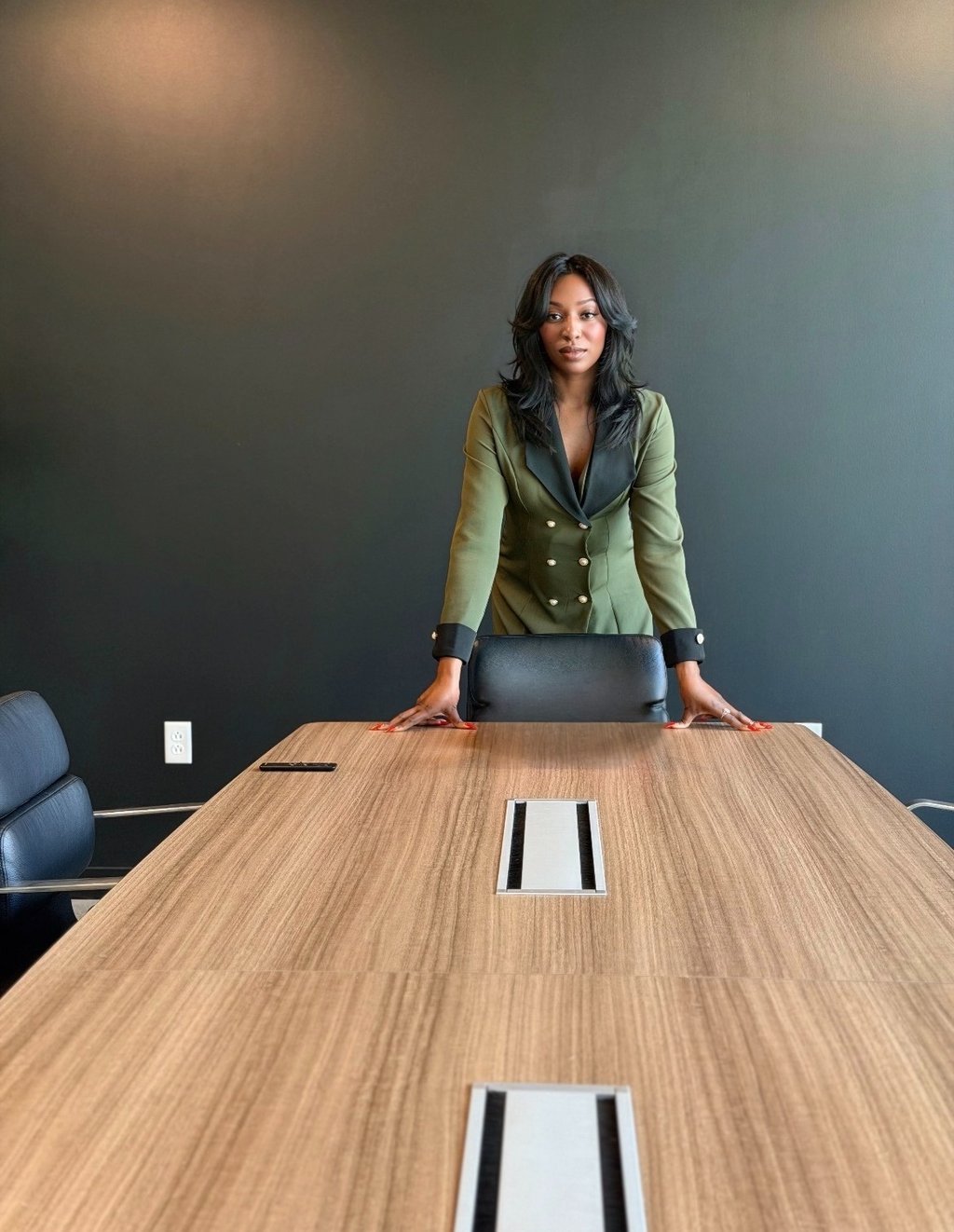 A confident professional woman in a green blazer standing at a large wooden boardroom table.