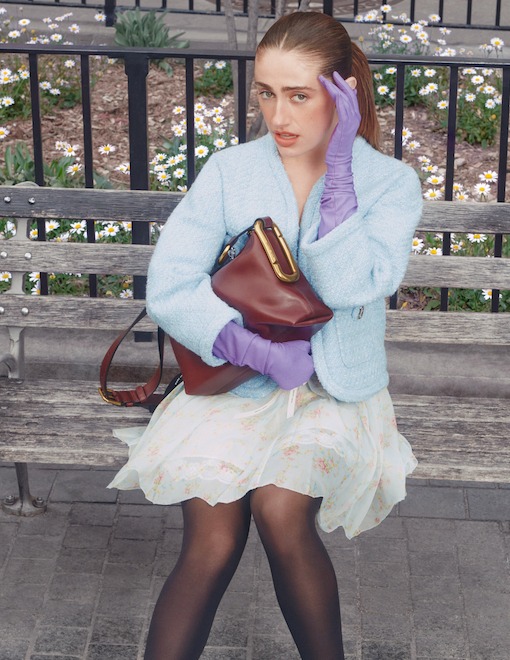 Fashionable woman in a blue tweed jacket and purple gloves holds a luxury leather bag on a park bench.