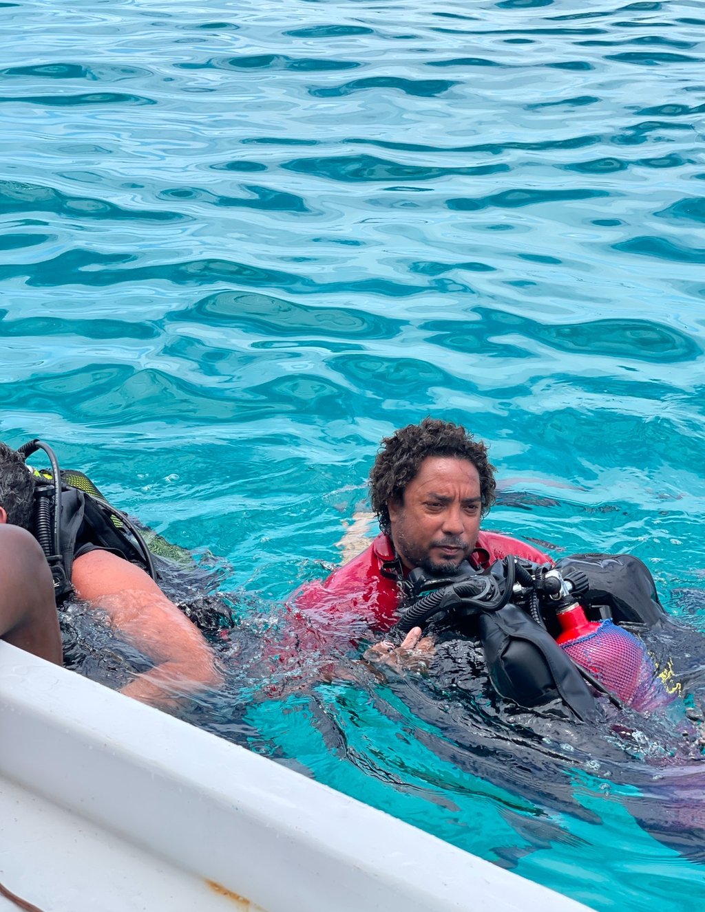 Scuba diver wearing a red shirt and full dive gear floating in clear blue tropical ocean water.