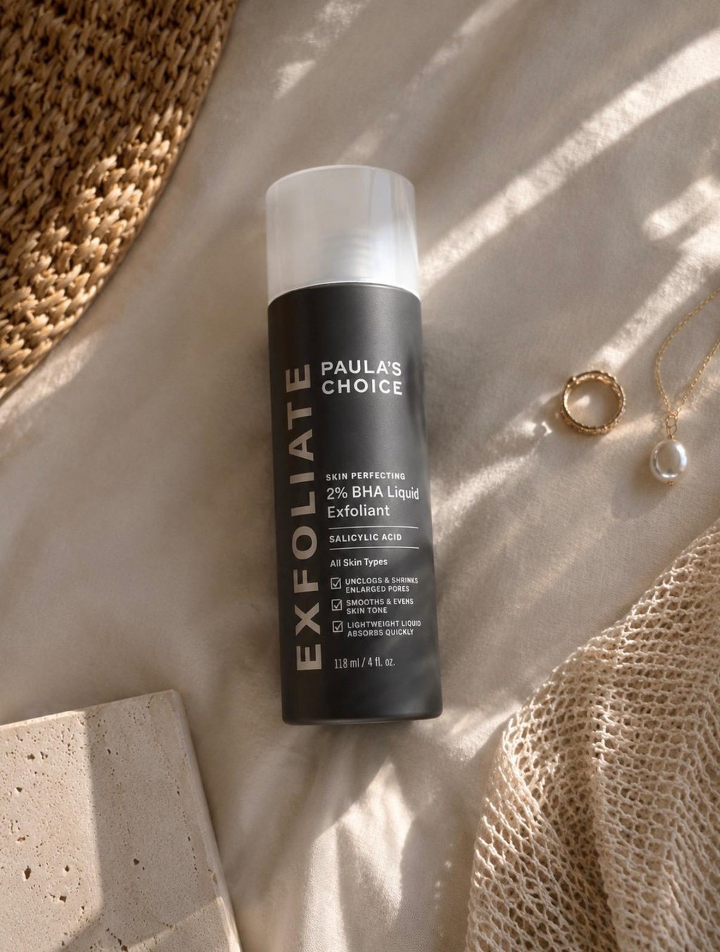 Paula’s Choice 2% BHA Exfoliant bottle styled on neutral fabric with gold jewelry