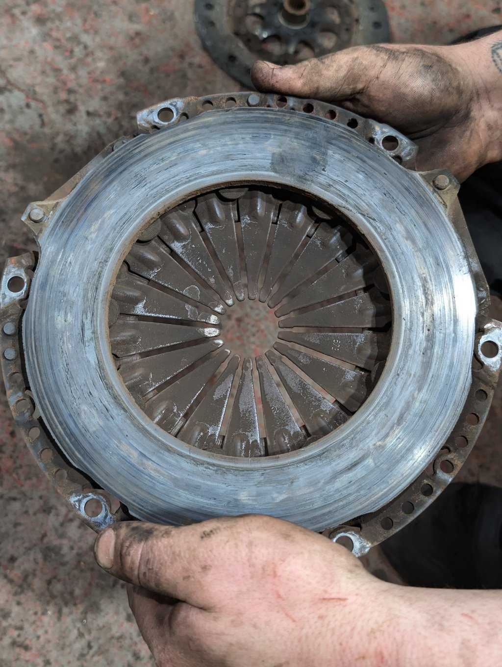 A mechanic holds a worn automotive clutch pressure plate showing signs of friction surface heat damage.