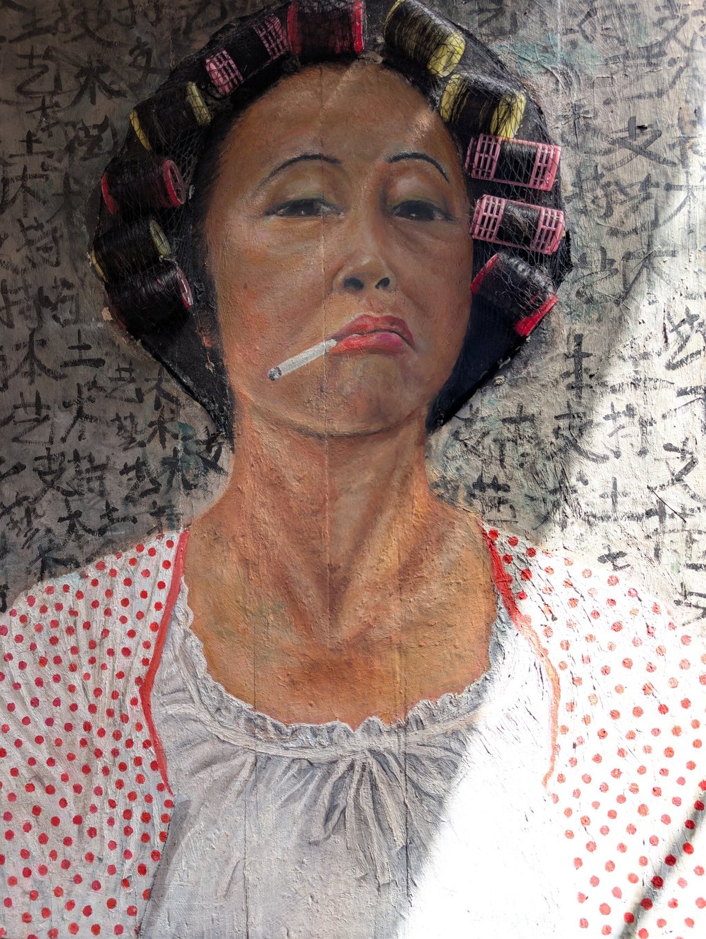 Street art mural of an Asian woman smoking a cigarette with hair rollers against a calligraphy background.