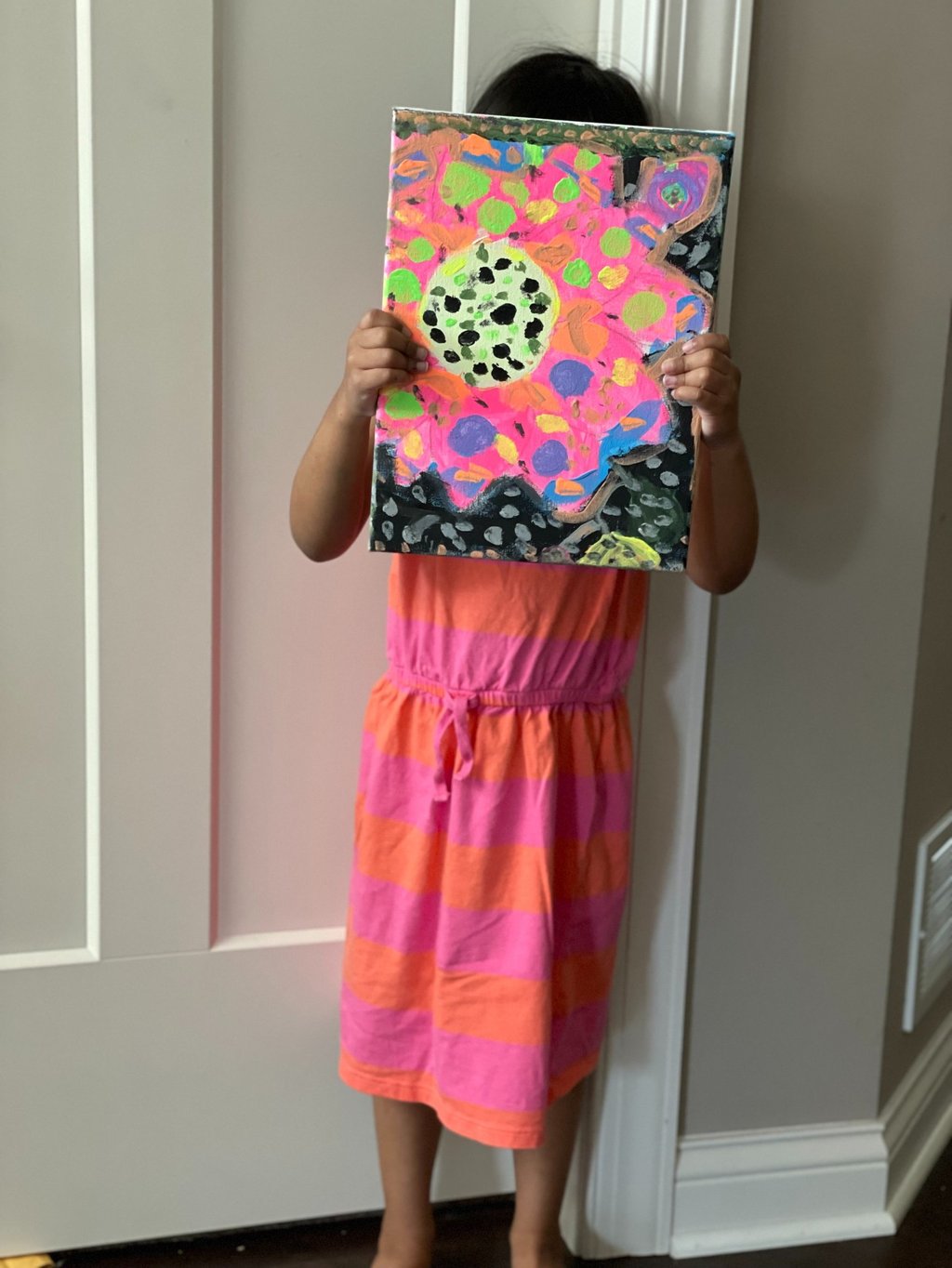 little girl with dark hair in a pink and orange dress holds colorful painting in front of her face