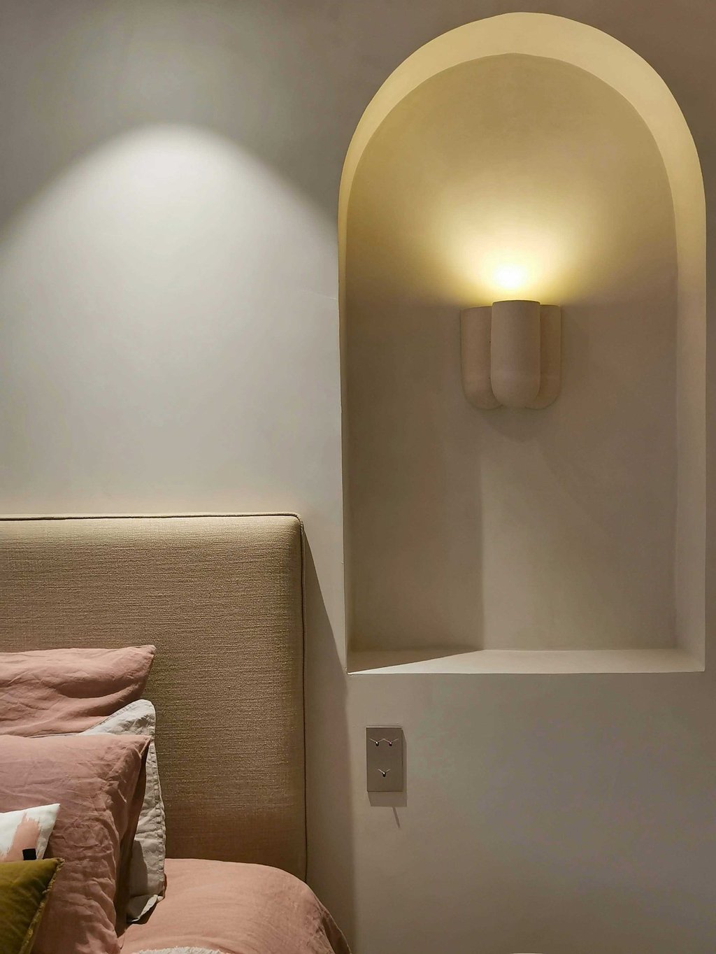 Modern minimalist bedroom featuring a beige upholstered headboard and a curved wall niche with a warm wall sconce.